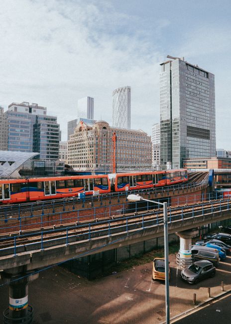 A modern urban scene featuring a bright orange train traveling on an elevated railway track against the backdrop of the Shadwell DLR area in the UK. Surrounding the railway, there are several contempo
