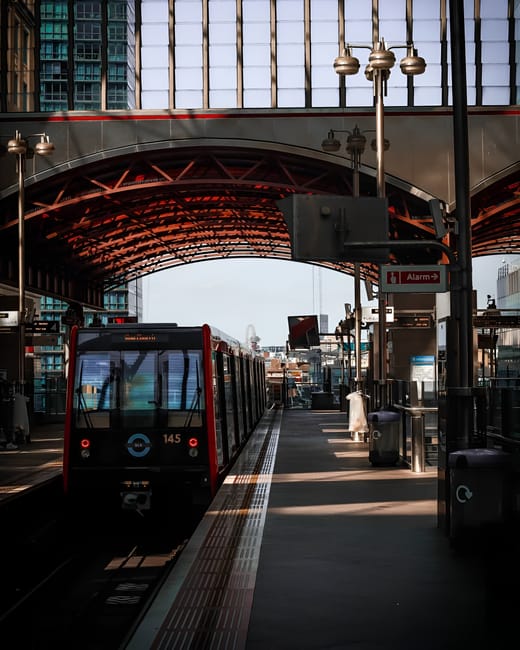 A modern urban scene featuring a bright orange train traveling on an elevated railway track against the backdrop of the Shadwell DLR area in the UK. Surrounding the railway, there are several contempo