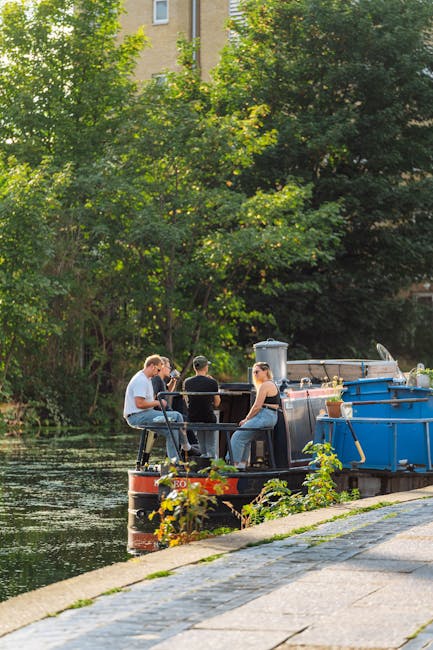 A quaint garden area situated alongside a narrow canal, featuring a variety of potted plants and small shrubs arranged along a black metal fence. In the canal, a dark green narrowboat with a small win